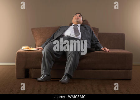 Homme d'obèses dans les vêtements officiels de dormir sur un canapé avec la plaque d'hamburgers et de frites Banque D'Images