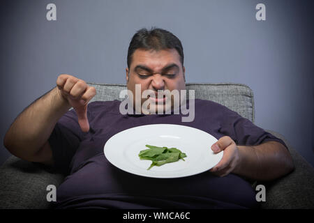 Obèses malheureux homme tenant une assiette avec quelques feuilles d'épinard et montrant un signe de pouce vers le bas Banque D'Images