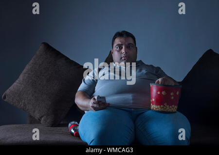 L'homme obèse à la maison à regarder la télévision assis sur le canapé avec le pop-corn à remous et bouteille de boisson Banque D'Images