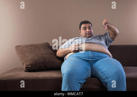 Obèses malheureux man sitting on sofa holding sa graisse du ventre d'une main et montrant Thumbs down signer avec d'autres Banque D'Images