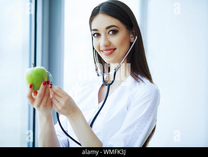 Santé et beauté. Médecin diététiste debout près de la fenêtre au bureau de lumière. Fille tient la pomme dans les mains et les sourices. Le médecin montre Apple Banque D'Images