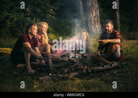 Groupe d'amis du camping en forêt. Deux couples de saucisses à frire sur des bâtons. Les jeunes à parler autour de feu de camp Banque D'Images