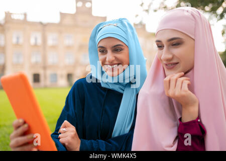 Student wearing hijab bleu holding orange tablet assis près de ami Banque D'Images