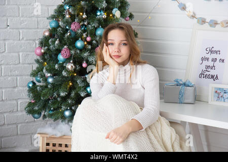 Portrait of smiling young woman looking at camera sur mur en brique blanche et l'arrière-plan de l'arbre de Noël Banque D'Images