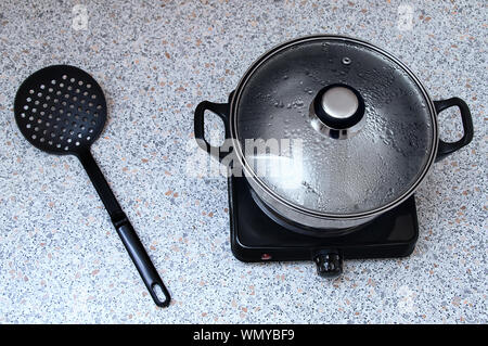 Casserole sur une petite cuisinière électrique avec un couvercle pour un couple. Sur la table est une cuillère écumoire en plastique noir. Vue de dessus. Banque D'Images