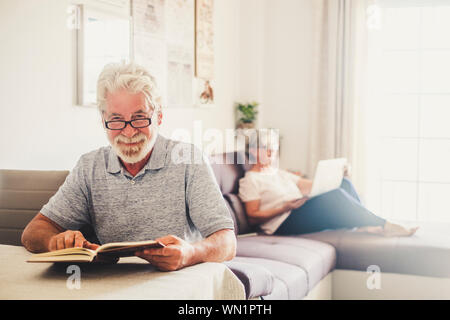 Senior man reading book que sa femme utilise un ordinateur portable dans la salle de séjour Banque D'Images