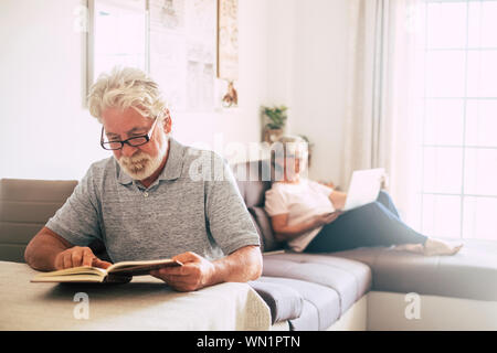 Senior man reading book que sa femme utilise un ordinateur portable dans la salle de séjour Banque D'Images