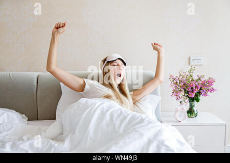 Happy young blonde woman en pyjama et bandeau de se réveiller, les bâillements et les étirements au lit, avoir une bonne journée. Lumière du matin dans la chambre. Détente Banque D'Images