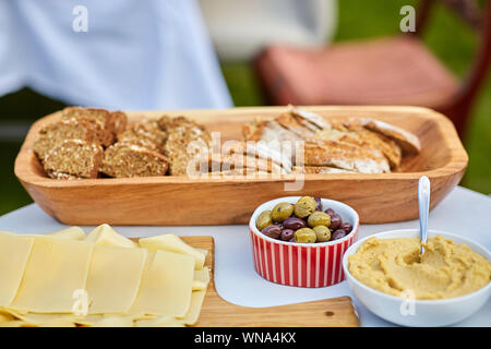 La nourriture servie sur la table lors de la garden party - plateau en bois avec des tranches de pain brun, olives vertes et noires dans le bol, l'hoummos et plateau de fromage. Banque D'Images