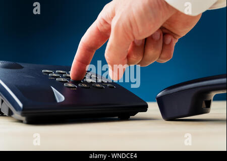 Gros plan du doigt masculin en appuyant sur un bouton sur le clavier du téléphone fixe sur un bureau. Sur fond bleu foncé. Banque D'Images