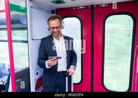 Businessman Standing On Train Film Streaming à partir d'un téléphone portable sur un casque sans fil Banque D'Images