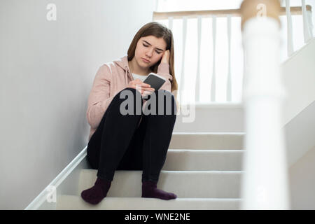 Adolescente malheureuse victime d'intimidation par message texte assis sur des escaliers à la maison Banque D'Images