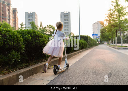 Petit enfant apprendre à conduire un scooter dans une ville sur la route de l'été ensoleillé jour. Mignon bambin fille en tutu jupe équitation un rouleau. Kid joue outdoor Banque D'Images