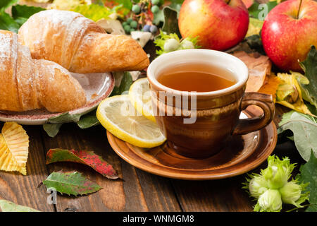 Tasse de thé au citron, croissant, l'automne feuilles d'automne, les fruits sur fond de bois. Accueil chaleureux, petit-déjeuner de style automne hygge, confort concept. Banque D'Images