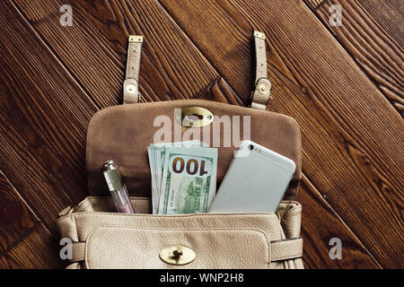 Vue de haut contenu de sac femme. Brillant à lèvres Mise en page, l'argent comptant, un téléphone mobile et des lunettes de soleil dans un sac en cuir marron sur fond de bois Banque D'Images