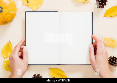 Mains tenant livre avec des pages vides sur les feuilles tombées, la citrouille et les cônes sur fond beige pastel. Composition d'automne, mise à plat, vue du dessus, les frais généraux Banque D'Images
