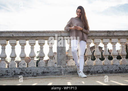 Femme souriante en utilisant son téléphone tout en s'appuyant sur balustrade en pierre Banque D'Images