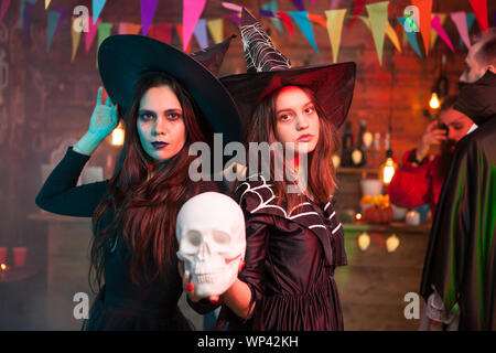 Deux belles filles en robes noires et chapeaux de sorcière tenir un crâne pour Halloween. Cheerful sorcières. Banque D'Images