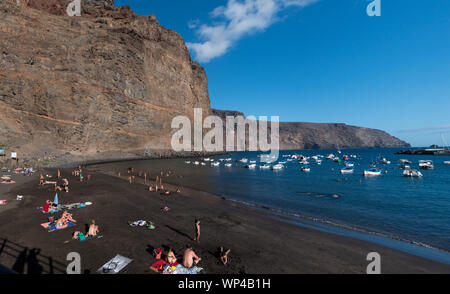 La Gomera, Valle Gran Rey, Espagne - 10 octobre 2018 : plage de sable noir dans le port avec des falaises spectaculaires, des baigneurs de soleil non identifiés, des bateaux et un ciel bleu Banque D'Images