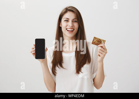 Smartphone acheté à crédit et fier. Studio shot of emotive happy young female student montrant votre carte bancaire et votre nouveau téléphone, se vante de nouveau pur Banque D'Images