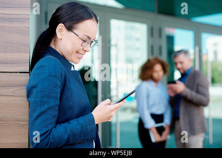 Happy young brunette femme défilant ou la lecture de message dans smartphone outdoors Banque D'Images