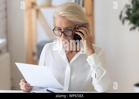 De graves senior woman talk on cellphone discussing paperwork Banque D'Images