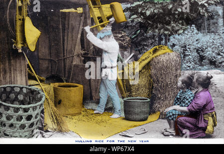 [ 1920 - Japon ] Riz japonais le décorticage des agriculteurs - un agriculteur est le décorticage du riz pendant que sa femme et son fils, tous deux portant un yukata, regarde. Cette carte postale est d'une série sur l'agriculture japonaise, appelé Farmer La vie au Japon. 20e siècle vintage carte postale. Banque D'Images