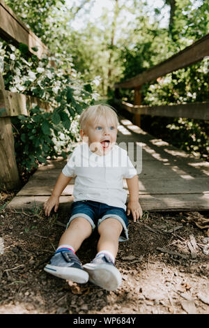 Mignon petit gamin Blond Bébé garçon enfant assis et de rire en face de pont de bois sur un ruisseau au parc dans la forêt en été Banque D'Images