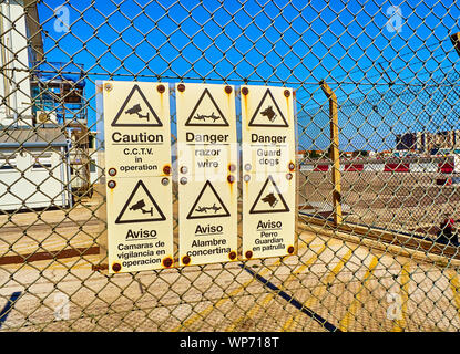 Danger et mise en garde en anglais et espagnol sur un mur de clôture de grillage métallique en acier. Banque D'Images