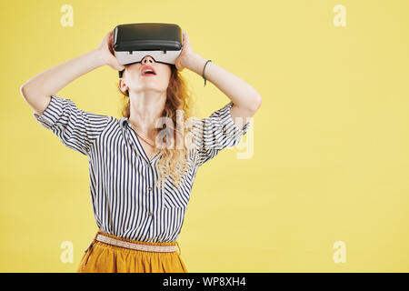 Jeune femme Schoked en chemise rayée debout contre fond jaune et l'exploration de la réalité virtuelle Banque D'Images