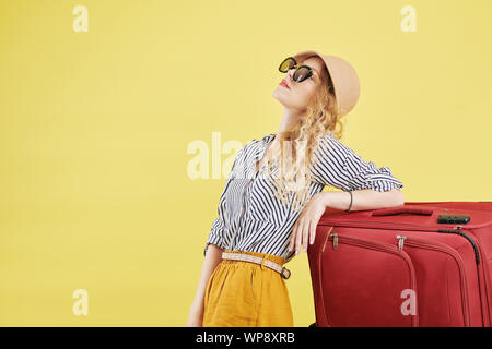 Jolie jeune femme dans des vêtements d'été, un chapeau et des lunettes de soleil s'appuyant sur big red suitcase, isolé sur fond jaune Banque D'Images