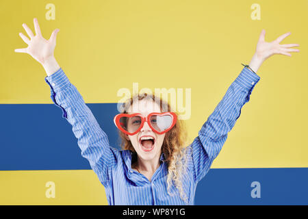 Jolie fille heureuse en forme de coeur dans l'augmentation des armes et lunettes criant à huis clos, isolé sur bleu et jaune. Banque D'Images