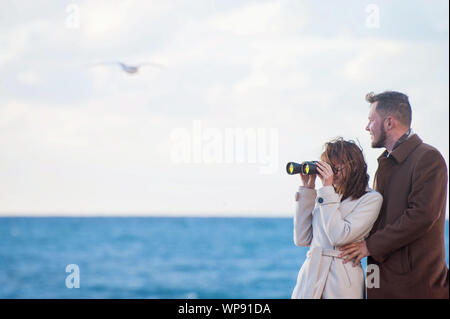 Beau jeune couple à la mode Homme et Femme au manteau binoculars proche mer avec horizon et flying seagull Banque D'Images