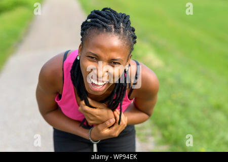 Jeune femme sportive africaine de flexion vers l'avant de rire à l'appareil photo alors qu'elle prend une pause de la course à pied sur un sentier rural Banque D'Images