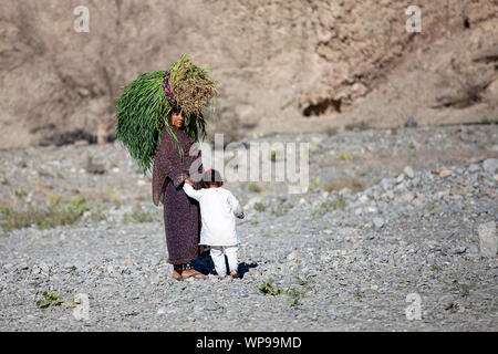 La femme est l'exécution sur la tête de grandes gerbes de foin et de tenir l'enfant dans un paysage aride en Oman Banque D'Images