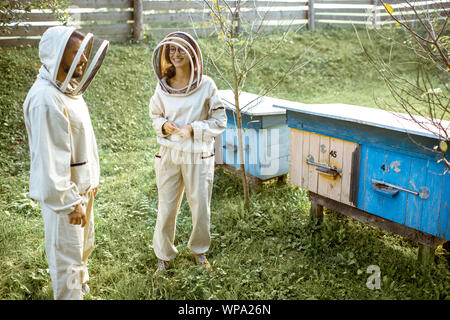 Deux jeunes en beekepers de travail uniformes de protection sur une petite ferme avec des ruches en bois rucher sur l'arrière-plan Banque D'Images