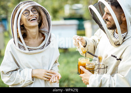 Libre de l'homme et la femme beekepers uniforme de protection en se tenant ensemble avec du miel dans le pot, dégustation de produits frais sur le rucher en plein air Banque D'Images