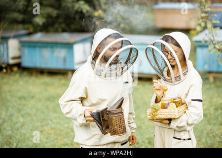 Deux beekepers en uniforme de protection avec beesmoker permanent et du miel, dégustation de produits frais sur le rucher Banque D'Images