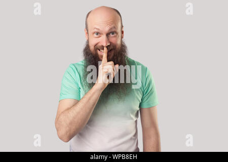 Chut, c'est secret. Portrait de dents smiley middle aged homme chauve avec longue barbe en vert clair t-shirt debout avec le doigt sur les lèvres et j'ai le sourire. Banque D'Images