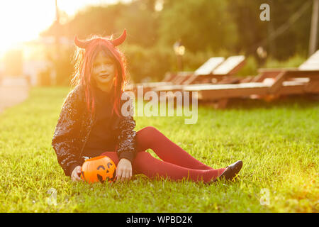 Portrait of cute little girl wearing costume Halloween assis sur pelouse verte avec panier citrouille et looking at camera, copy space Banque D'Images