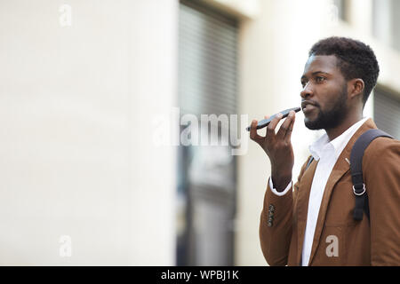 Vue de côté de l'homme afro-américain contemporain portrait message vocal d'enregistrement via smartphone tout en marchant dans la rue ville, copy space Banque D'Images