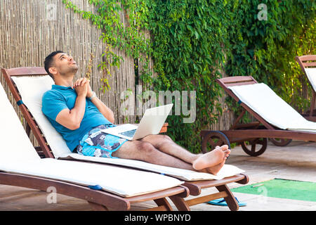 Portrait de la belle prière des jeunes adultes barbus dans l'homme Tee-shirt bleu et un short confortable avec transats sur ordinateur portable sur la piscine et le plaisir d'il Banque D'Images