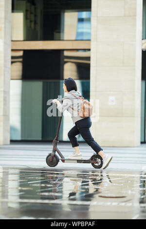 Vue latérale sur toute la longueur de Young Asian woman riding scooter électrique rapide en ville rue, copy space Banque D'Images