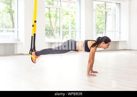 Side view portrait of young woman wearing musculaire haut noir et leggings debout sur plank position Utilisez des sangles de remise en forme, faire des tractions pendant que les jambes han Banque D'Images