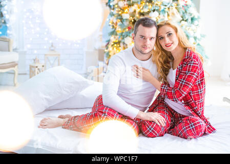 Loving couple mari et femme en Pyjama à carreaux sont assis près du Nouvel An des arbres par la cheminée et s'étreindre. L'amour et une famille heureuse. Banque D'Images