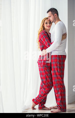 L'amour heureux couple mari et femme en Pyjama à carreaux sont à proximité d'une grande fenêtre et s'étreindre. Fenêtres en plastique, une chaleureuse maison confortable Banque D'Images
