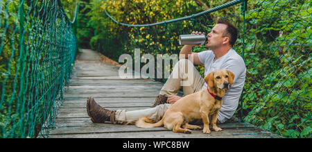 Randonneur avec petit chien jaune reposant sur le pont suspendu en bois dans la forêt et l'eau potable Banque D'Images