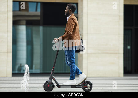 Vue de côté toute la longueur de l'afro-américaine moderne businessman riding scooter électrique lors des trajets de travailler en ville, copy space Banque D'Images