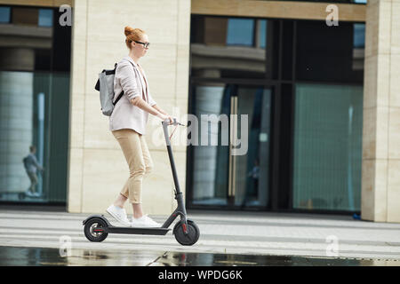 Vue de côté toute la longueur de jeune femme moderne riding scooter électrique lors des trajets de travailler en ville, copy space Banque D'Images