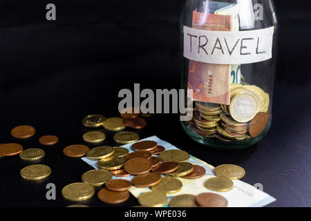 Transparent en verre pot étiqueté voyage manuscrite. Billets et pièces à l'intérieur et l'extérieur du pot. Fond noir isolé. Banque D'Images